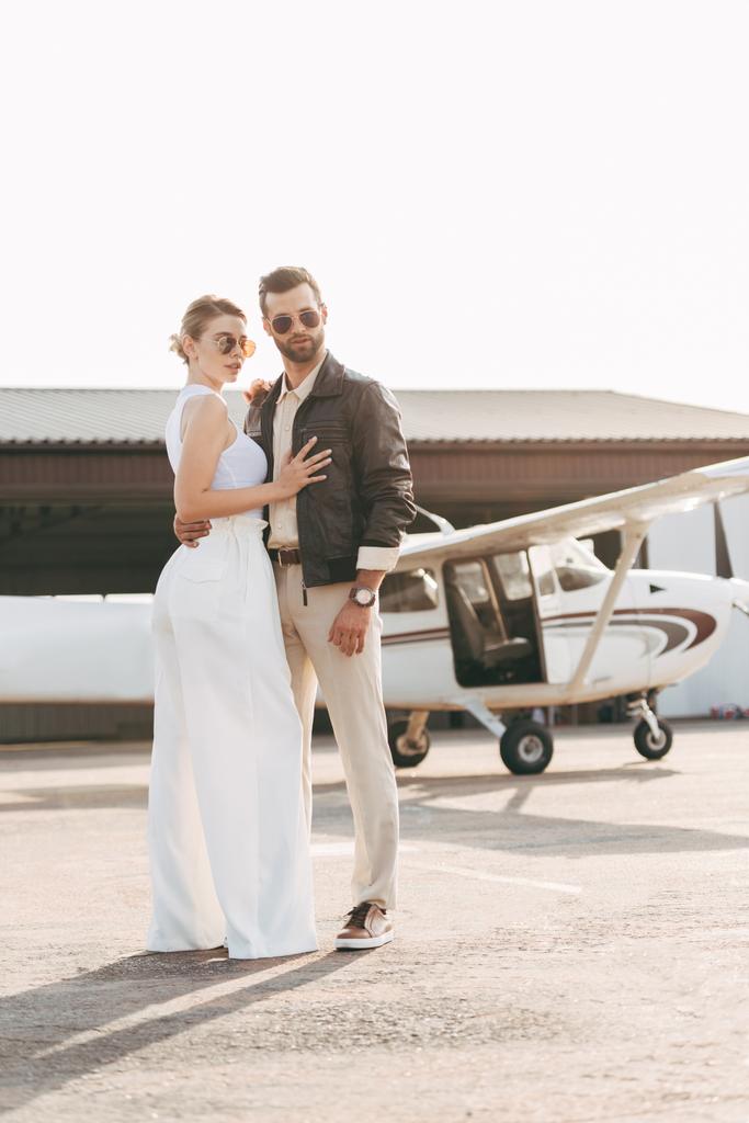 jovem bonito em jaqueta de couro e óculos de sol abraçando namorada atraente perto do avião
 - Foto, Imagem