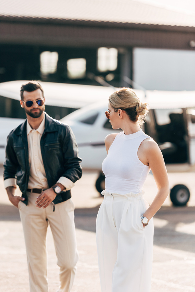 selektiver Fokus eines modischen jungen Paares mit Sonnenbrille, das in Flugzeugnähe spaziert  - Foto, Bild