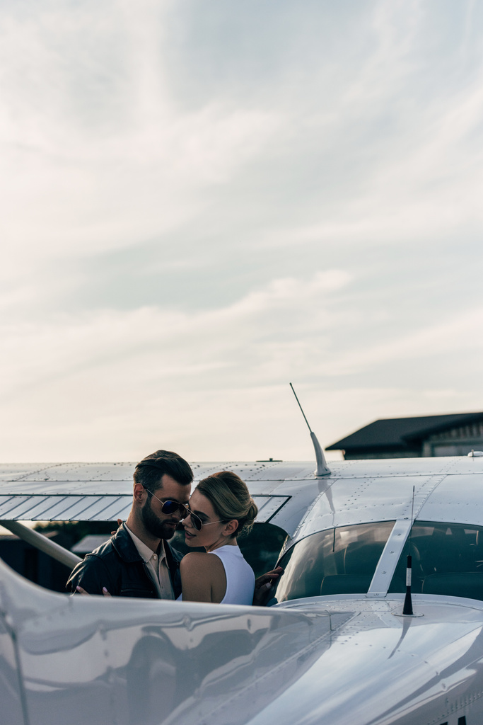 ernster gutaussehender Mann in Lederjacke und Sonnenbrille umarmt attraktive Freundin in der Nähe von Flugzeug  - Foto, Bild