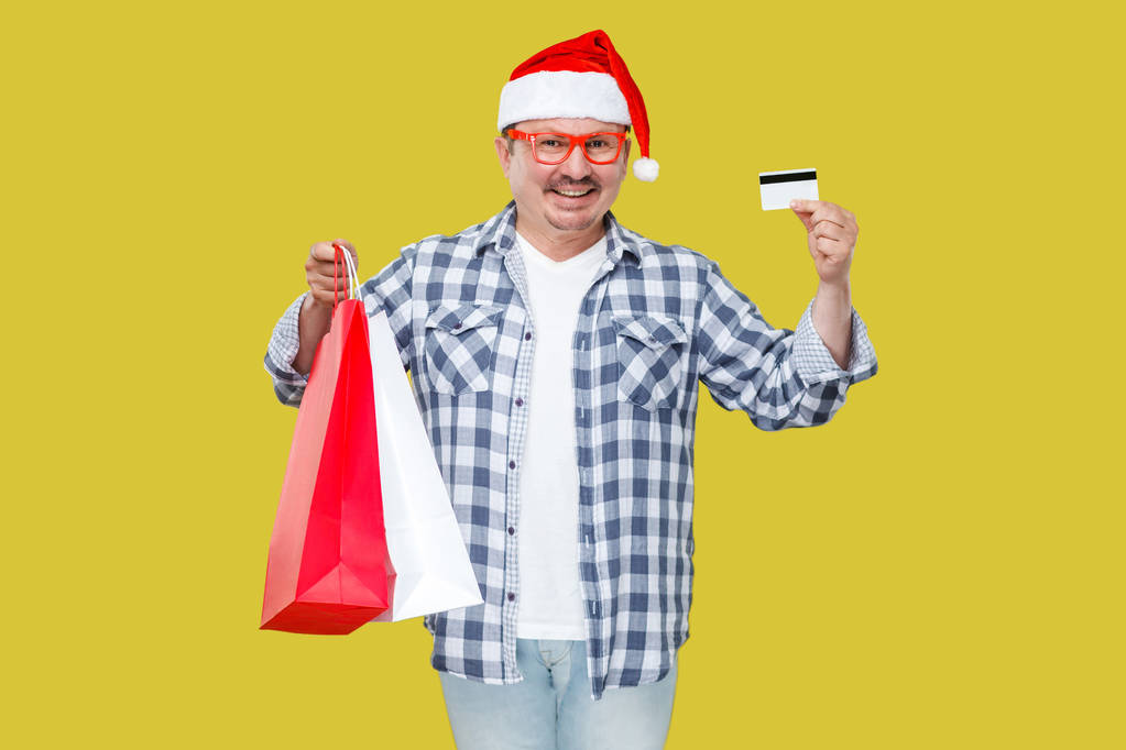 Hombre feliz en gorra roja sosteniendo bolsas de regalo de compras y tarjeta de crédito sobre fondo amarillo
 - Foto, Imagen