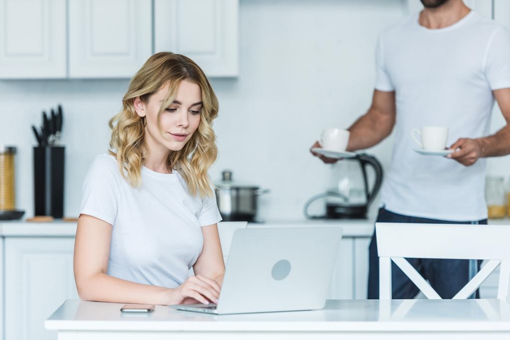 attraente sorridente ragazza utilizzando il computer portatile mentre il fidanzato in possesso di tazze di caffè dietro
 - Foto, immagini