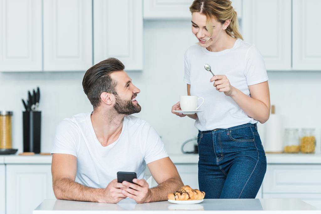 felice uomo barbuto utilizzando smartphone e guardando sorridente fidanzata in possesso di una tazza di caffè al mattino
 - Foto, immagini