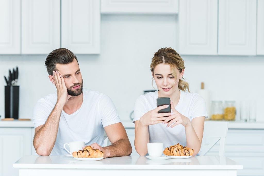 uomo annoiato guardando sorridente fidanzata utilizzando smartphone durante la colazione
 - Foto, immagini