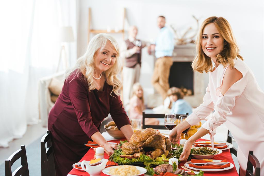 gelukkig jong en senior vrouwen ten dienste van thanksgiving day tabel terwijl mannen en kinderen die permanent op de achtergrond wazig - Foto, afbeelding