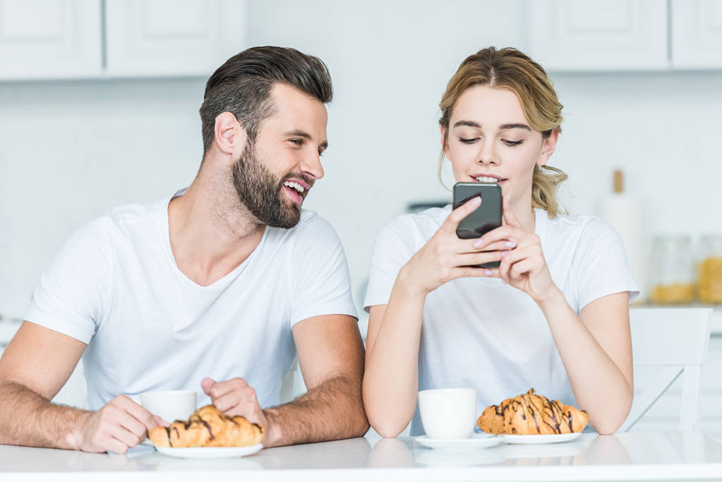 Улыбающийся молодой человек смотрит на подругу с помощью смартфона во время завтрака
 - Фото, изображение