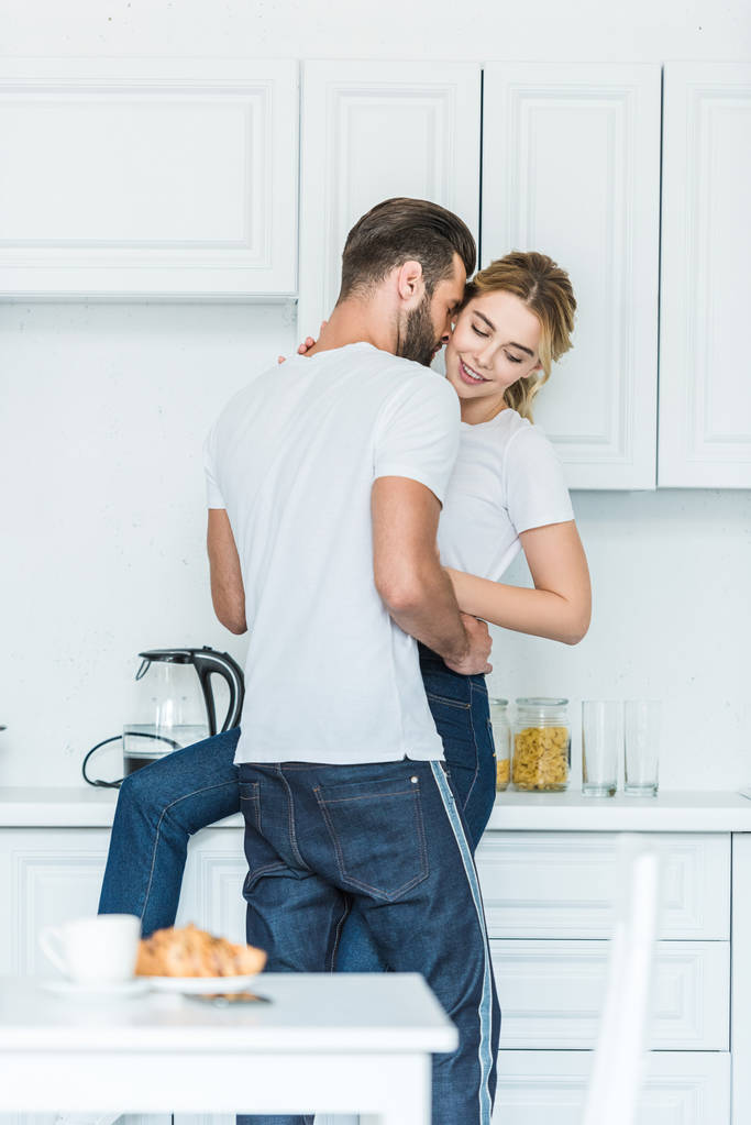 handsome young man hugging attractive smiling girlfriend in kitchen - Photo, Image