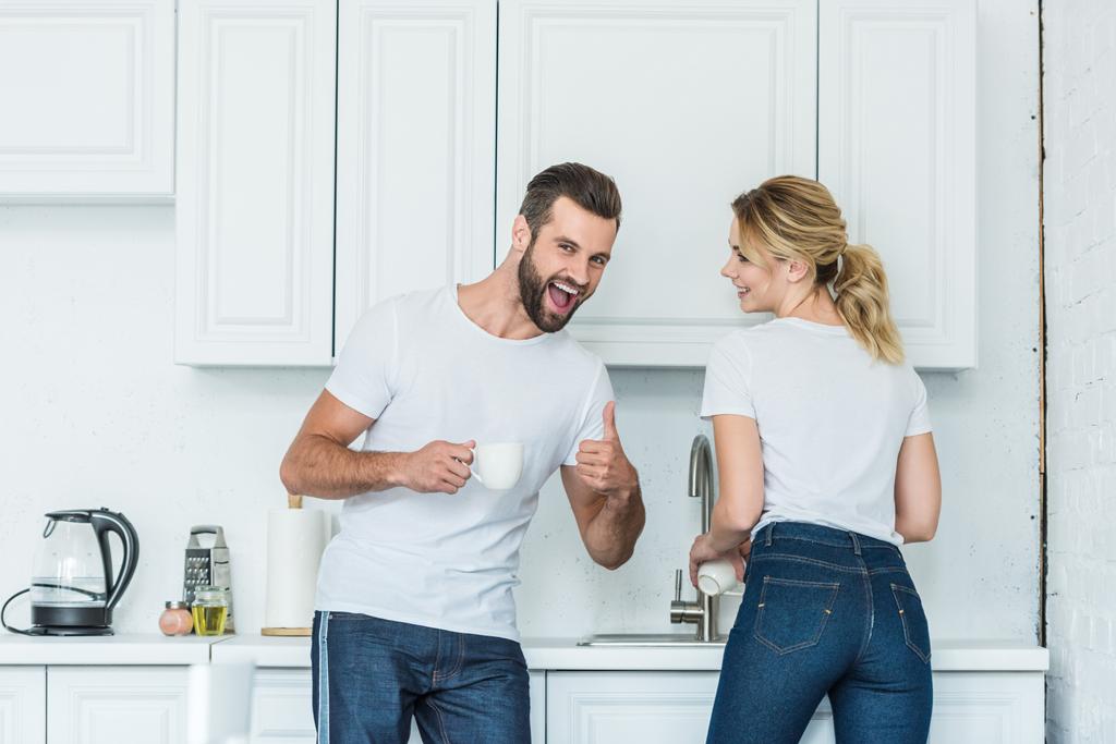 fröhliches junges Paar lacht gemeinsam in der Küche, Mann zeigt Daumen hoch und lächelt in die Kamera - Foto, Bild