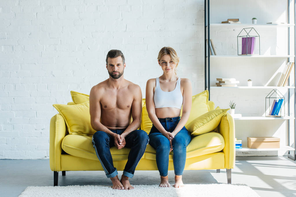 красивая молодая пара в джинсах сидит на диване и смотрит в камеру
 - Фото, изображение