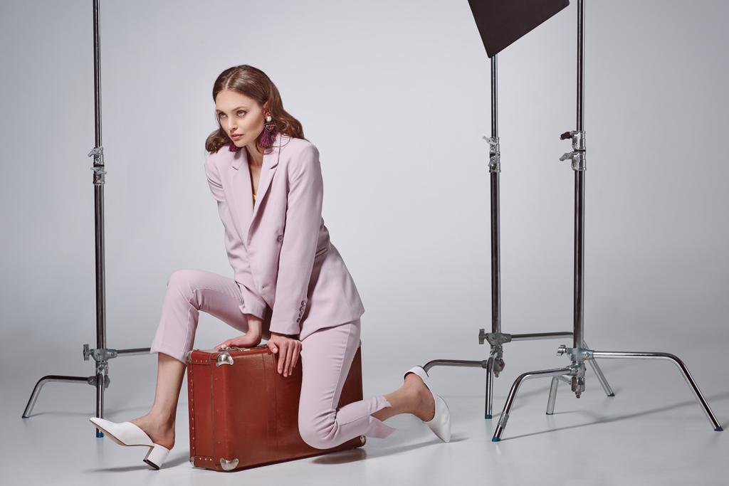 beautiful stylish woman in pink suit sitting on suitcase and looking away in recording studio - Photo, Image