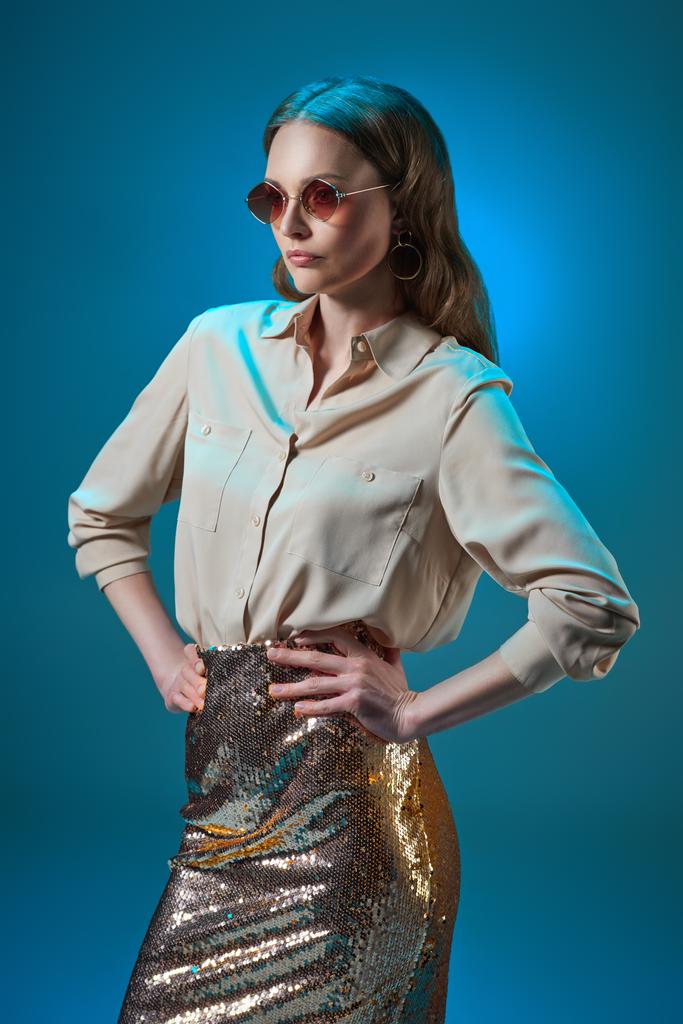 beautiful woman in fashionable golden glittering skirt and sunglasses posing with hands on waist isolated on blue - Photo, Image