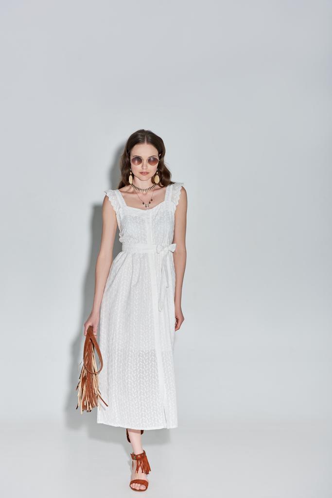 volle lengte weergave van mooie vrouw in stijlvolle witte jurk en zonnebril handtas te houden en lopen op grijs - Foto, afbeelding