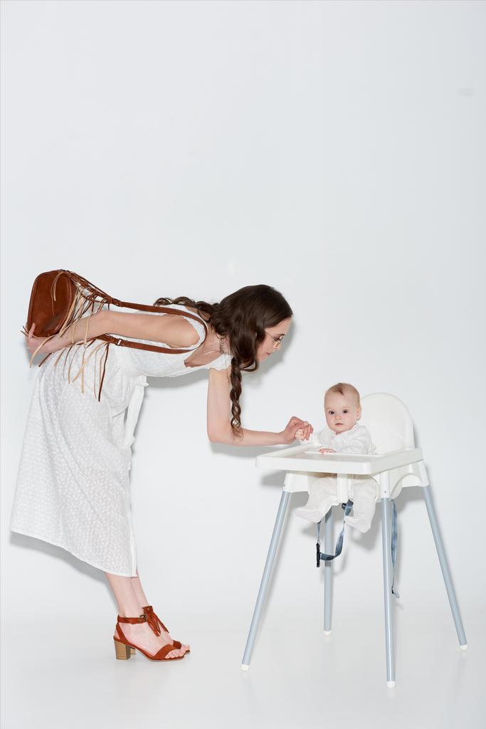 bela mulher elegante olhando adorável criança filha sentada em cadeira alta no cinza
 - Foto, Imagem