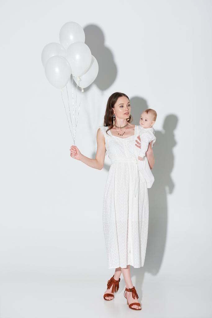 όμορφη γυναίκα στο κομψό λευκό φόρεμα που μεταφέρουν αξιολάτρευτο κοριτσάκι και κρατώντας μάτσο μπαλόνια σε γκρι  - Φωτογραφία, εικόνα