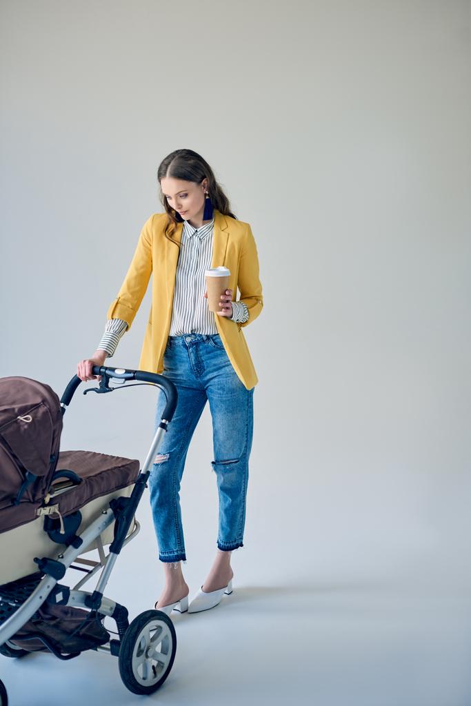 trendy jonge vrouw houden wegwerp koffiekopje en baby wandelwagen op grijs    - Foto, afbeelding