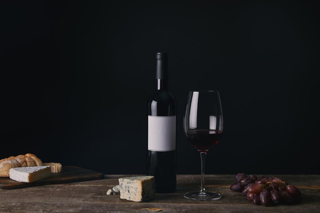 Flasche Wein mit leerem Etikett, Glas Rotwein, Käse und Trauben auf Holztisch - Foto, Bild