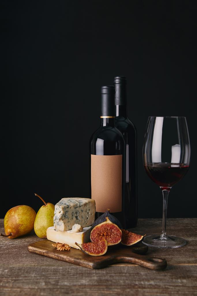 close-up view of bottles and glass of red wine, delicious cheese, figs and pears on wooden table - Photo, Image