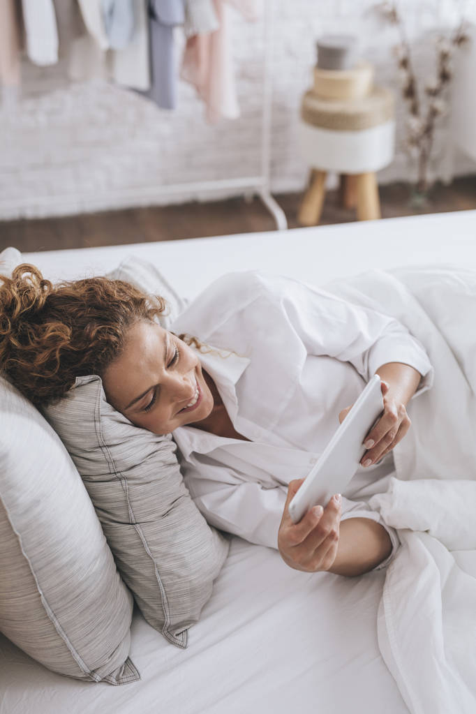 Mujer caucásica bastante sonriente acostada en su cama y leyendo en una tableta
. - Foto, imagen