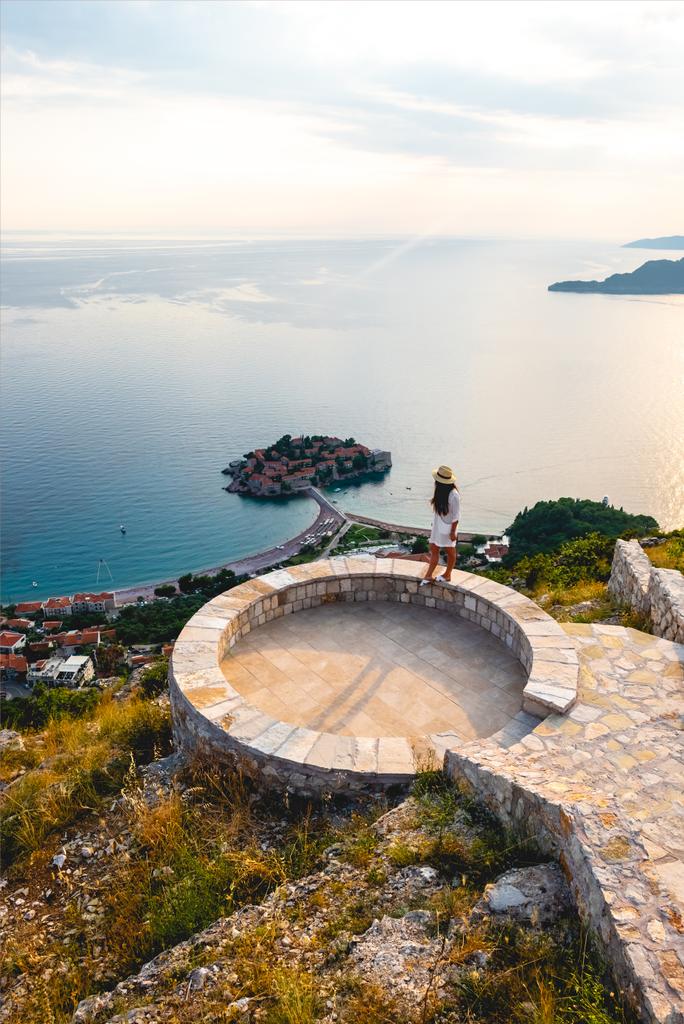 vista de alto ângulo da mulher em pé no miradouro perto da ilha de Santo Estefão no mar Adriático, Budva, Montenegro
 - Foto, Imagem