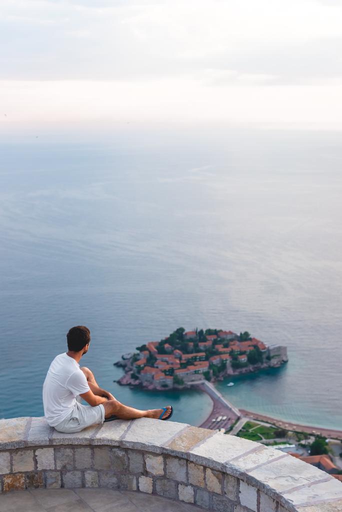 Mann sitzt auf einem Aussichtspunkt und blickt auf die Insel Sweti Stefan mit Hotelresort in der Adria, Budva, Montenegro - Foto, Bild
