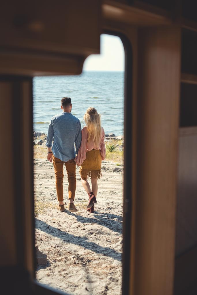 back view of hippie couple holding hands and walking near sea, from trailer door - Photo, Image