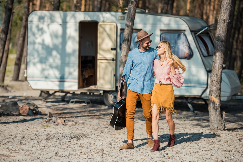beautiful couple hugging and walking with acoustic guitar near campervan in forest - Photo, Image