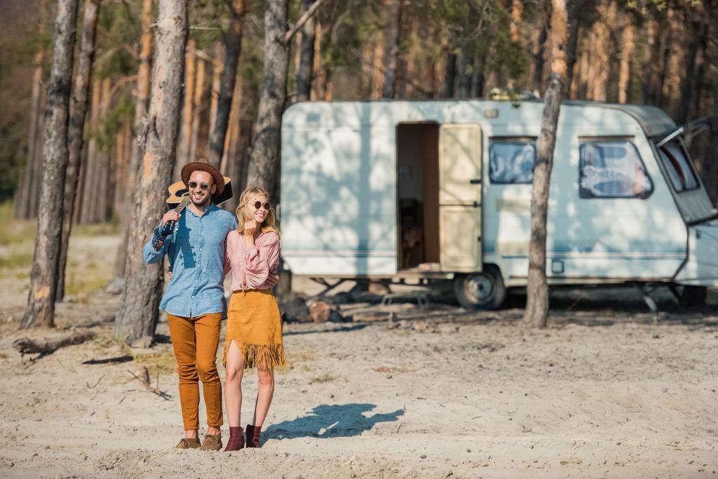 junges Hippie-Paar umarmt und steht mit Gitarre neben Wohnmobil im Wald - Foto, Bild