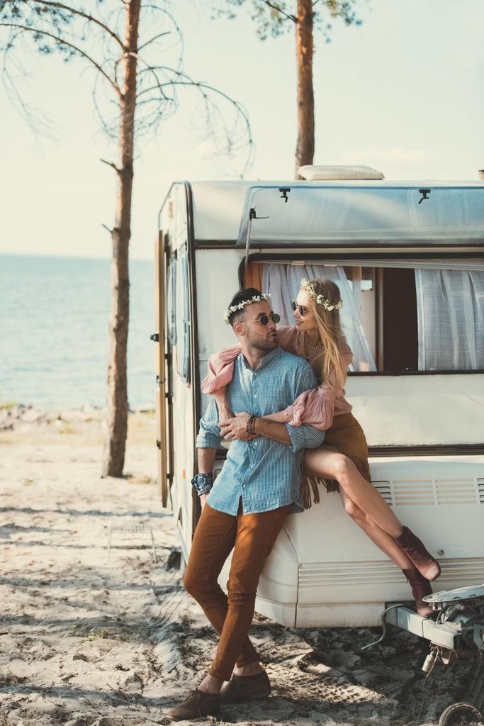 hippie couple in wreaths and sunglasses embracing and sitting on trailer   - Photo, Image