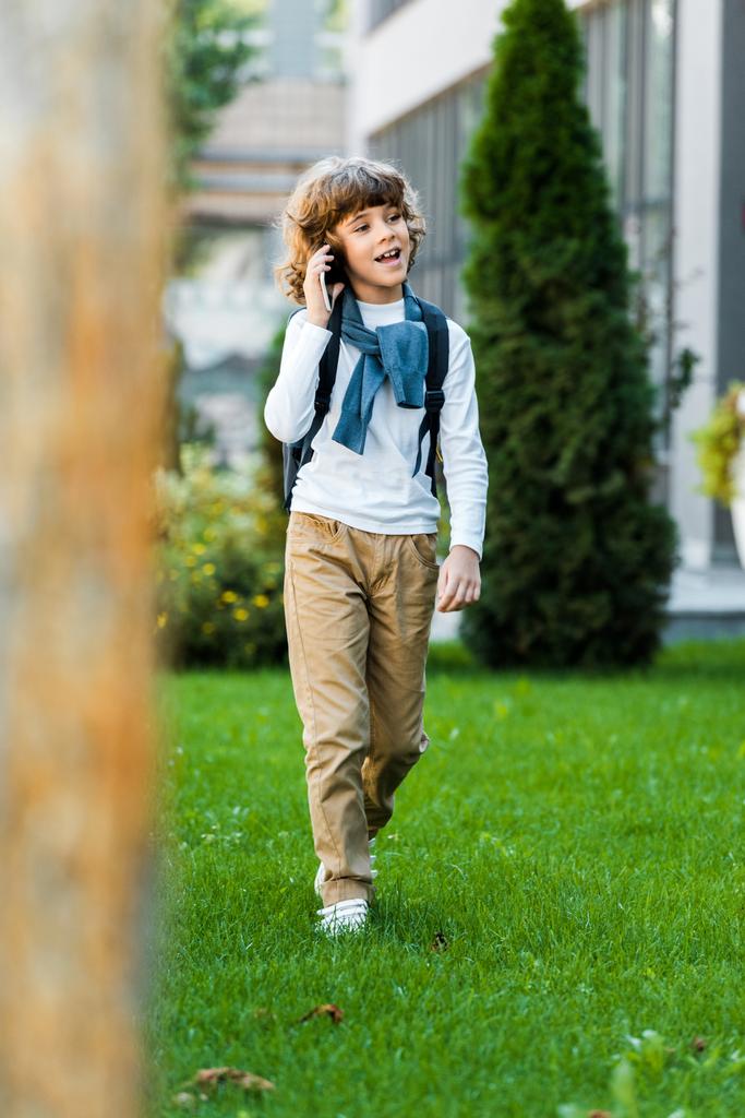 selective focus of smiling boy with backpack talking by smartphone and walking on green lawn - Photo, Image