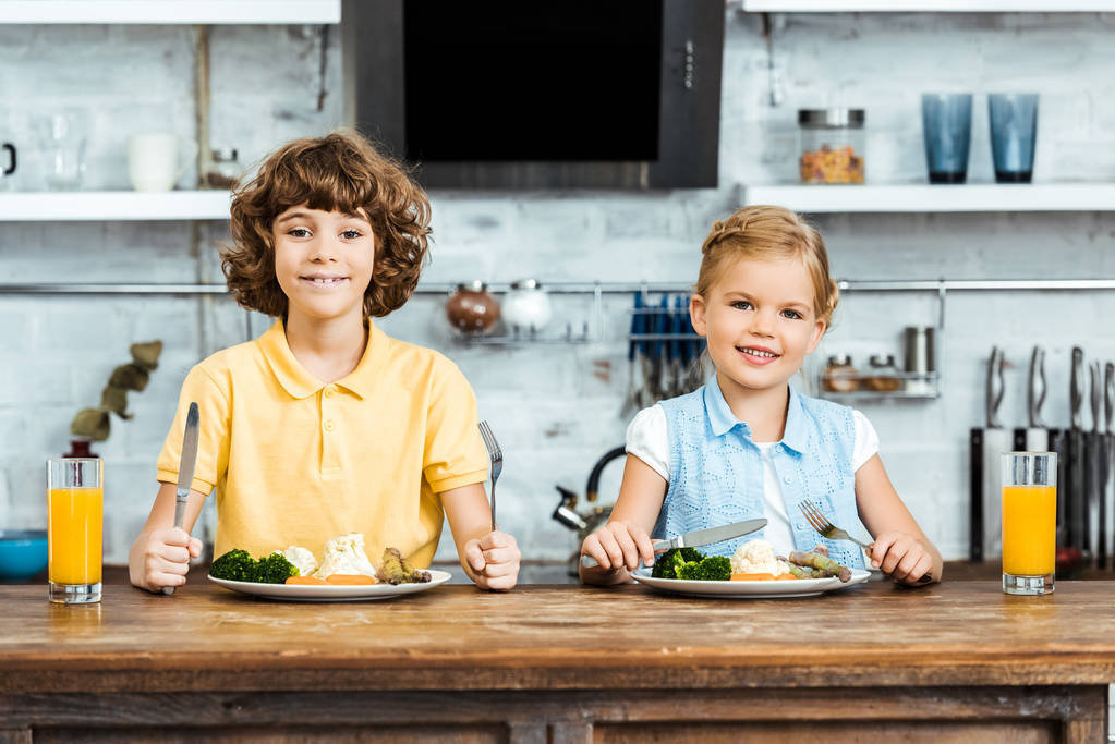 adorable kids eating vegetables and smiling at camera in kitchen - Photo, Image