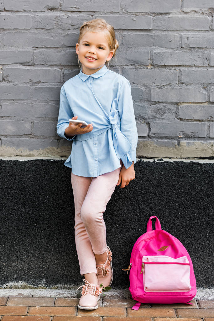 petite écolière mignonne utilisant un smartphone et souriant à la caméra tout en se penchant sur le mur de briques avec sac à dos rose
 - Photo, image