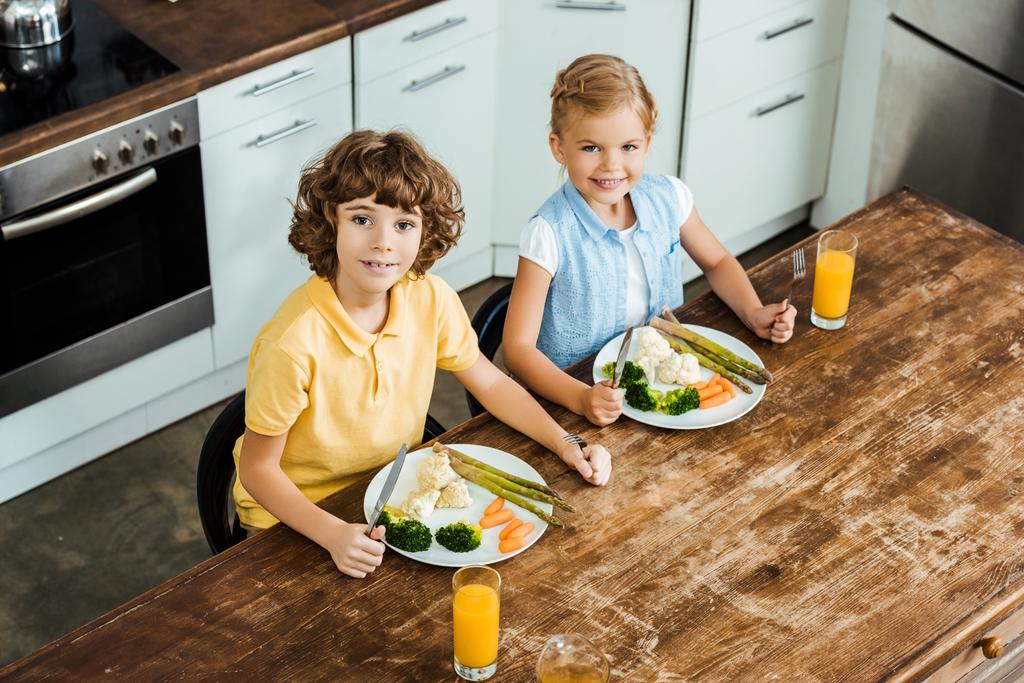 vista de ángulo alto de lindos niños felices comiendo verduras saludables y sonriendo a la cámara
 - Foto, imagen