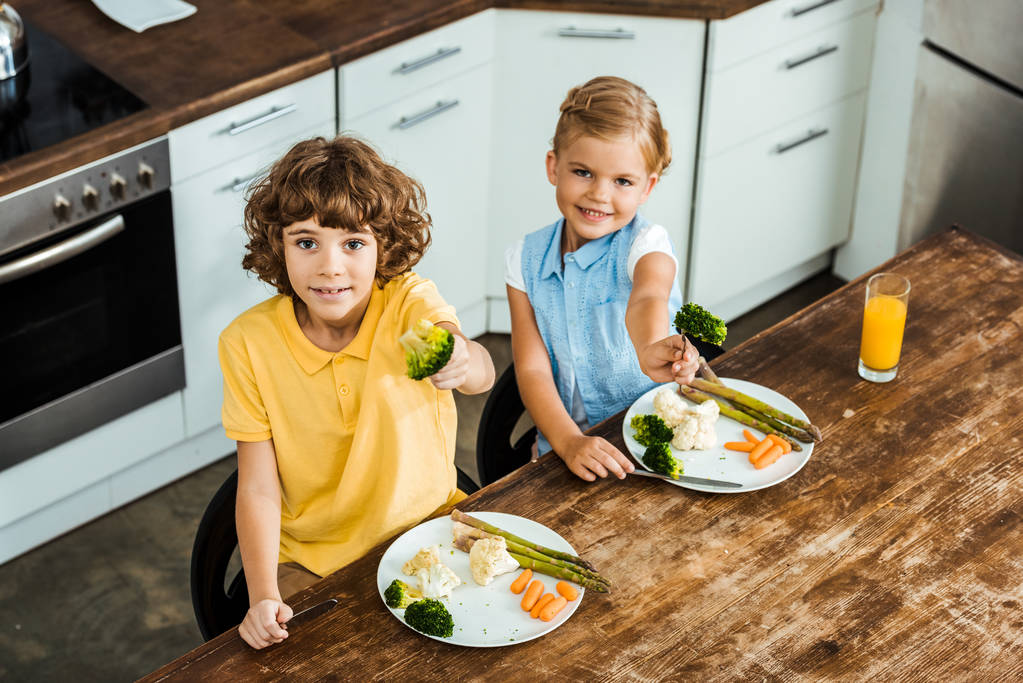 pohled z vysokého úhlu šťastný roztomilý dětí drží vidlice s brokolicí a usmívá se na kameru - Fotografie, Obrázek