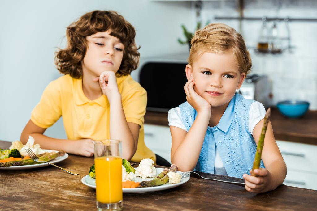 verveelde kinderen kijken naar asperges zittend aan tafel met groenten op platen - Foto, afbeelding