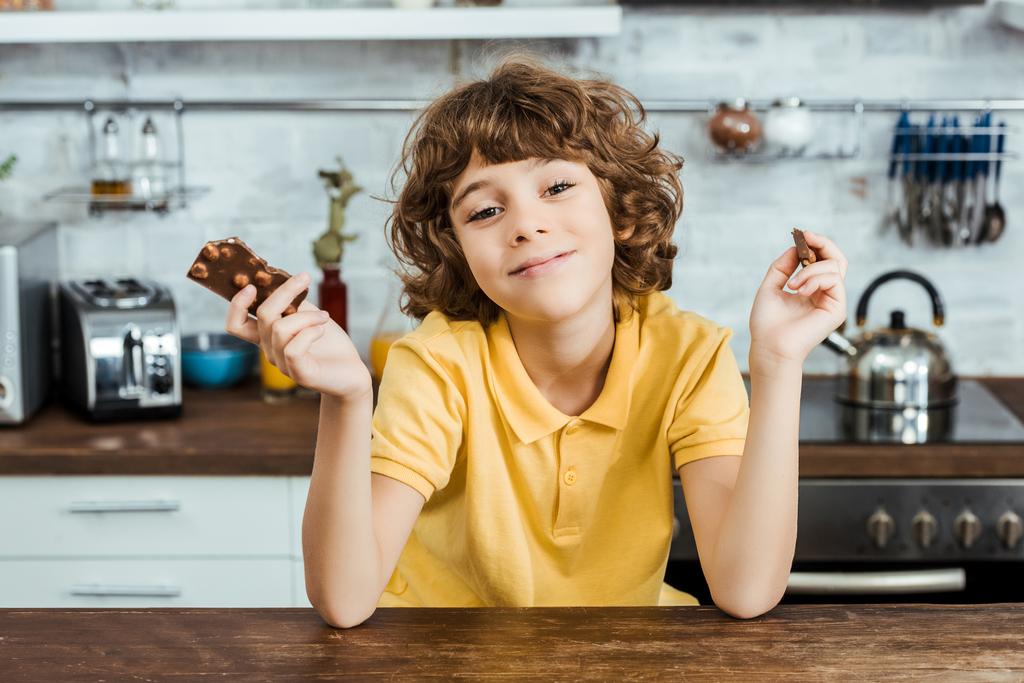 αξιολάτρευτο παιδί ευτυχισμένη κρατώντας κομμάτια από τη λαχταριστή σοκολάτα και να χαμογελά στη φωτογραφική μηχανή  - Φωτογραφία, εικόνα