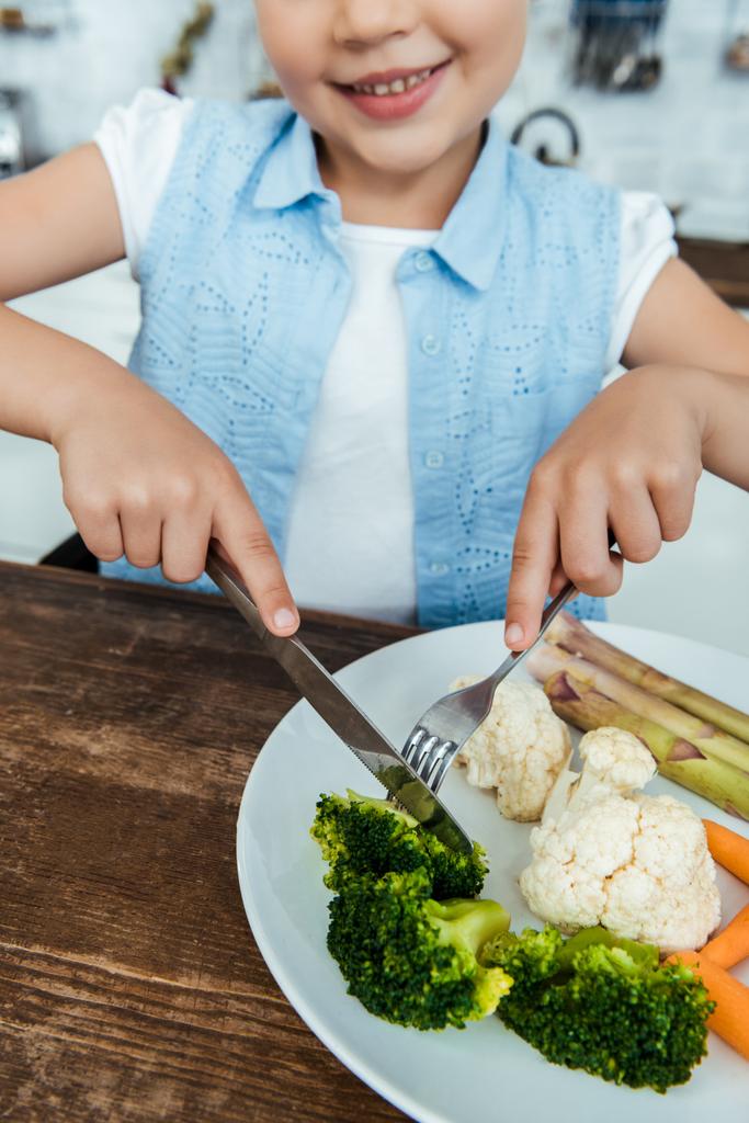 abgeschnittene Aufnahme eines lächelnden Kindes, das Gabel mit Messer hält und Brokkoli isst - Foto, Bild