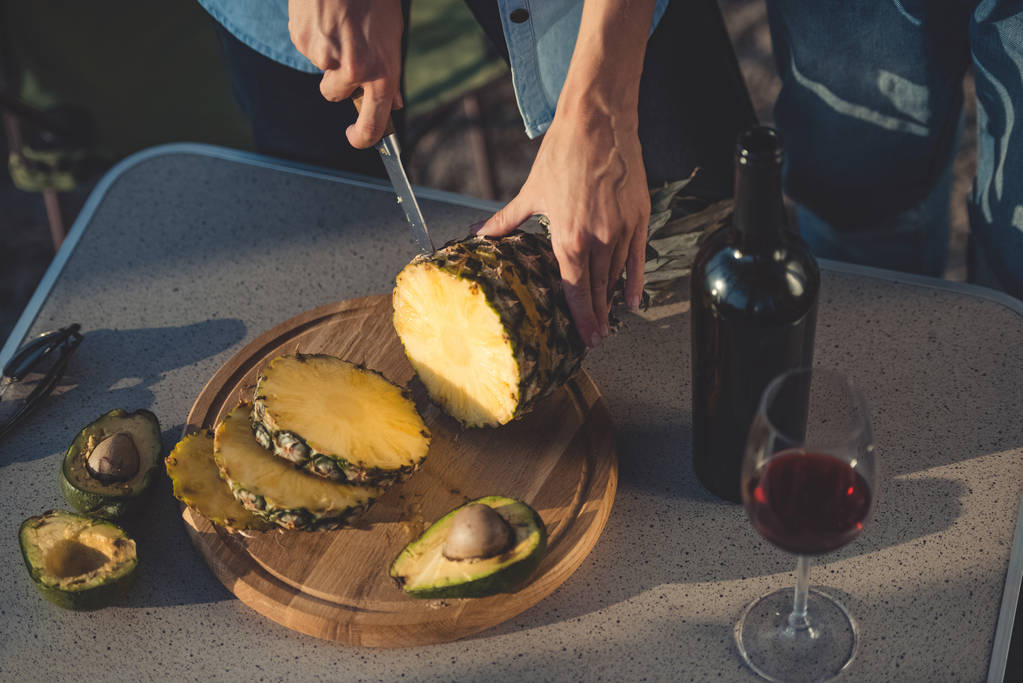 cropped view of woman cutting pineapple and avocados on wooden board near bottle of red wine - Photo, Image