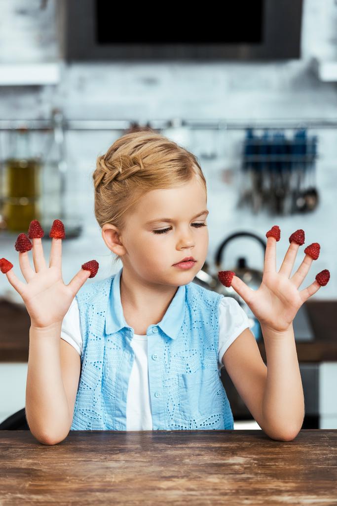 criança pequena bonito sentado à mesa e olhando para framboesas frescas maduras nos dedos
  - Foto, Imagem