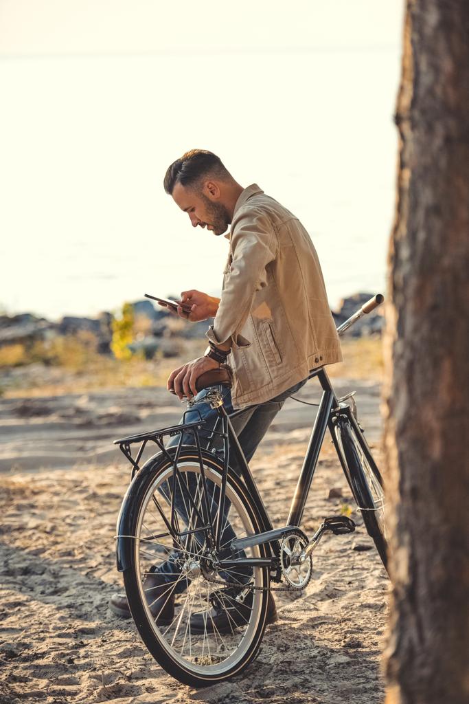 joven hombre usando gadget y apoyándose en bicicleta cerca del mar
 - Foto, imagen