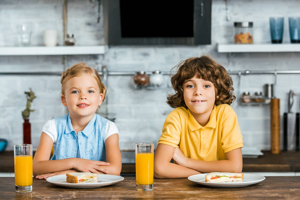 aranyos boldog gyerekek fényképezőgép gyümölcslé és ízletes szendvicseket, szemüveg asztalnál ülve mosolyogva - Fotó, kép