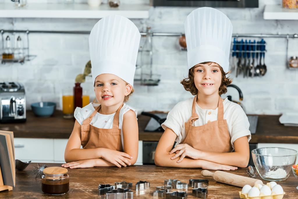 エプロンやシェフの帽子が一緒にキッチンで料理をしながらカメラに笑顔のかわいい幸せな子供たち - 写真・画像