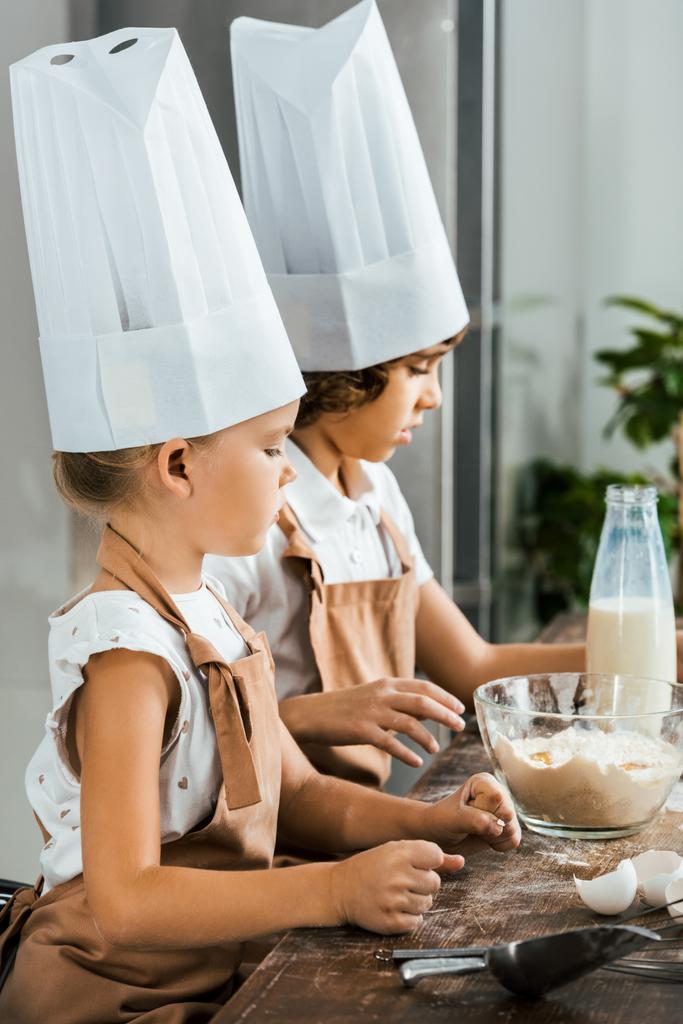 Zijaanzicht van schattige kinderen in chef mutsen en schorten voorbereiding van deeg voor cookies in de keuken - Foto, afbeelding