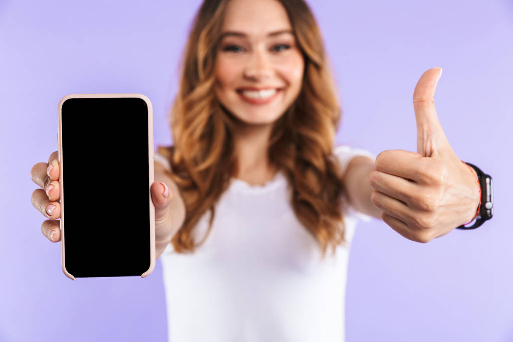 Κοντινό πλάνο του μια ενθουσιασμένος νεαρό κορίτσι στέκεται απομονωμένο πάνω από μοβ φόντο, προβάλλονται κενή οθόνη κινητό τηλέφωνο, δείχνει τους αντίχειρες επάνω - Φωτογραφία, εικόνα