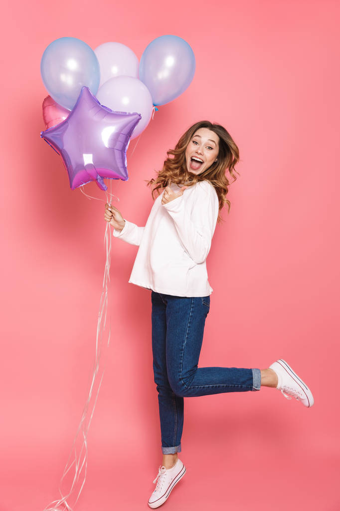 Ganzkörperporträt einer fröhlichen jungen Frau, die ein Bündel Luftballons in der Hand hält, während sie isoliert über rosa Hintergrund springt und wegzeigt - Foto, Bild