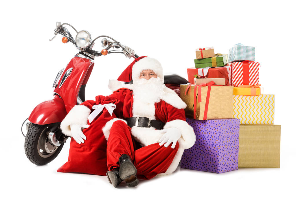 усталый Санта Клаус сидит на полу с винтажным красным скутером, мешок и подарочные коробки изолированы на белом
 - Фото, изображение