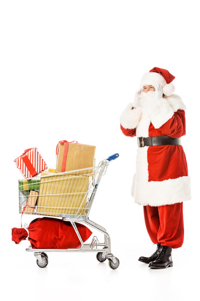 Seitenansicht des Weihnachtsmannes mit Einkaufswagen voller Geschenkboxen, die isoliert auf weißem Grund wegschauen - Foto, Bild
