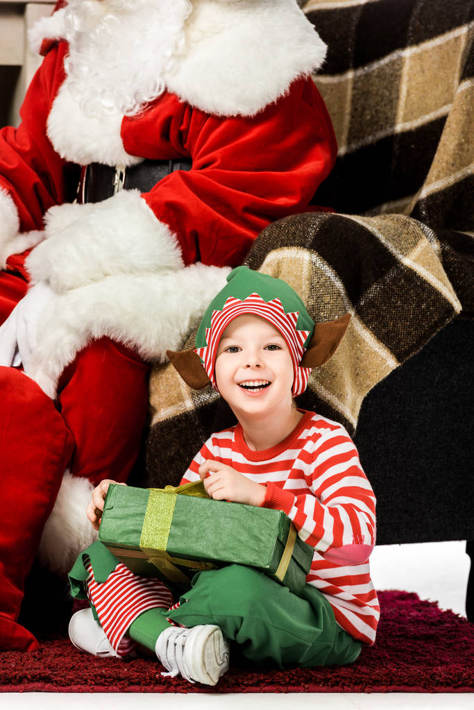 χαρούμενος μικρό παιδί κάθεται στο χαλί με Χριστουγεννιάτικο δώρο κοντά σε santa σε πολυθρόνα - Φωτογραφία, εικόνα