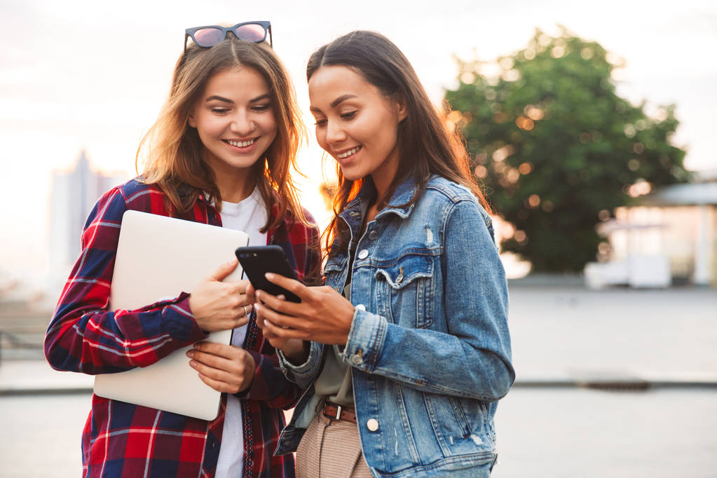 Две счастливые юные подруги стоят вместе на городской улице, держа в руках ноутбук и мобильный телефон, разговаривая
 - Фото, изображение