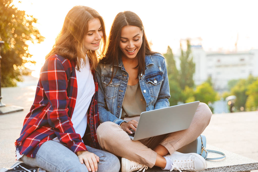 Deux jeunes filles heureuses amies assises ensemble dans la rue de la ville, utilisant un ordinateur portable, parlant
 - Photo, image