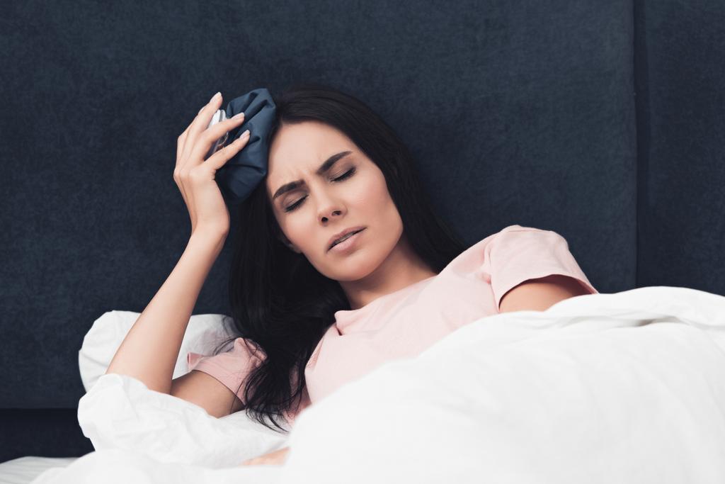enfermo joven mujer sosteniendo bolsa de hielo en la cabeza mientras está acostado en la cama
 - Foto, Imagen