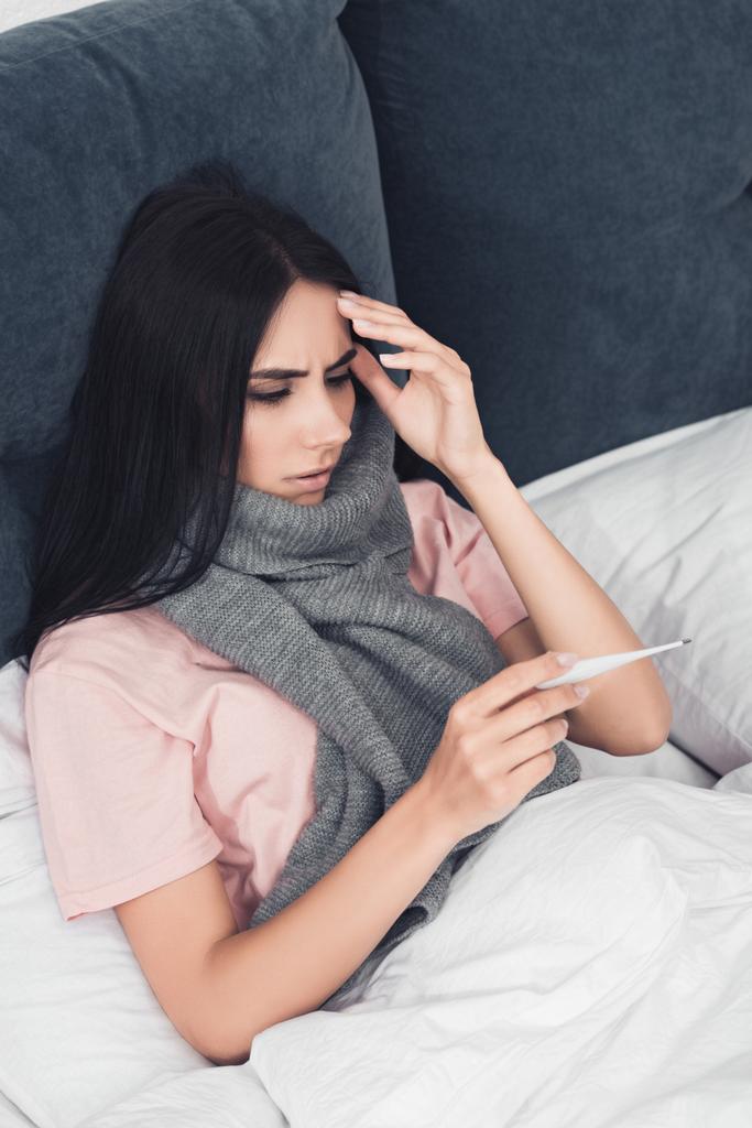 zieke jonge vrouw in sjaal elektronische thermometer kijken terwijl liggend in bed - Foto, afbeelding